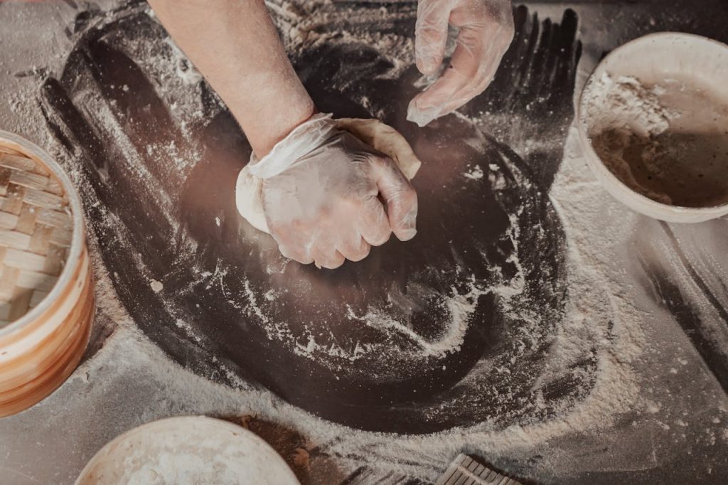 Hands in gloves kneading dough on a floured table with bowls and baking tools nearby.